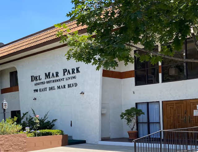 Front entrance of the Del Mar Park assisted retirement living building with signage, wooden double doors, windows, and surrounding landscaping.