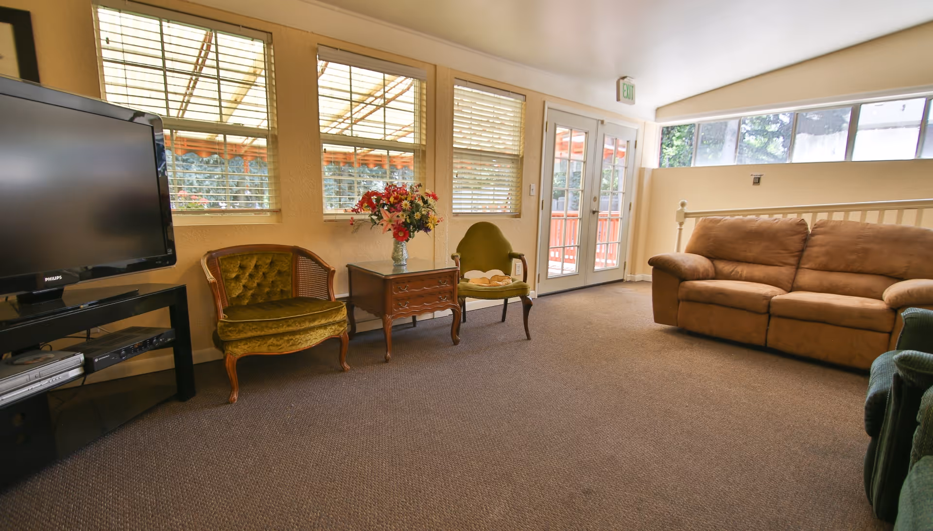 A cozy living room with a brown sofa, two vintage green armchairs, a wooden side table with a vase of flowers, a flat-screen TV on a black stand, and large windows letting in natural light. There is a door leading outside and carpeted flooring.
