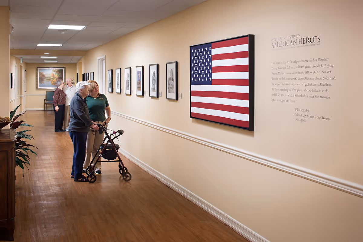 Residents and a staff member stand in a senior living hallway looking at framed photos and a large American flag display on the wall.