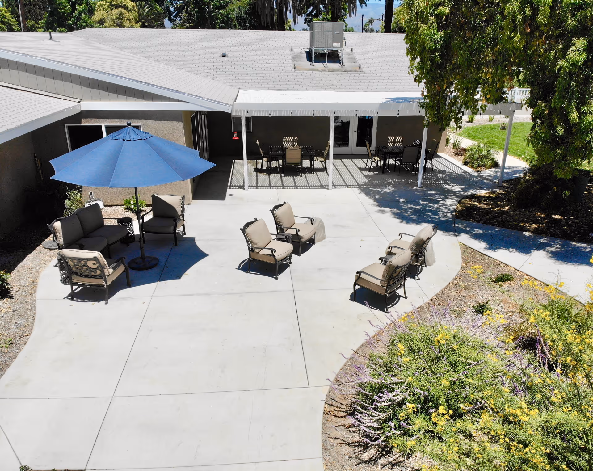 Outdoor patio area with several cushioned chairs arranged in small groups on a concrete surface. There is a large blue umbrella providing shade over one seating area. The patio is adjacent to a building with a covered section containing tables and chairs. Surrounding the patio are landscaped areas with plants and trees.