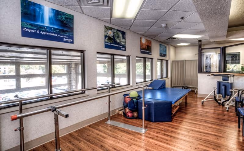 A rehabilitation therapy room with wooden flooring, parallel bars for walking exercises, a blue padded therapy table, exercise balls, and various therapy equipment. The room has large windows letting in natural light and motivational posters on the walls.