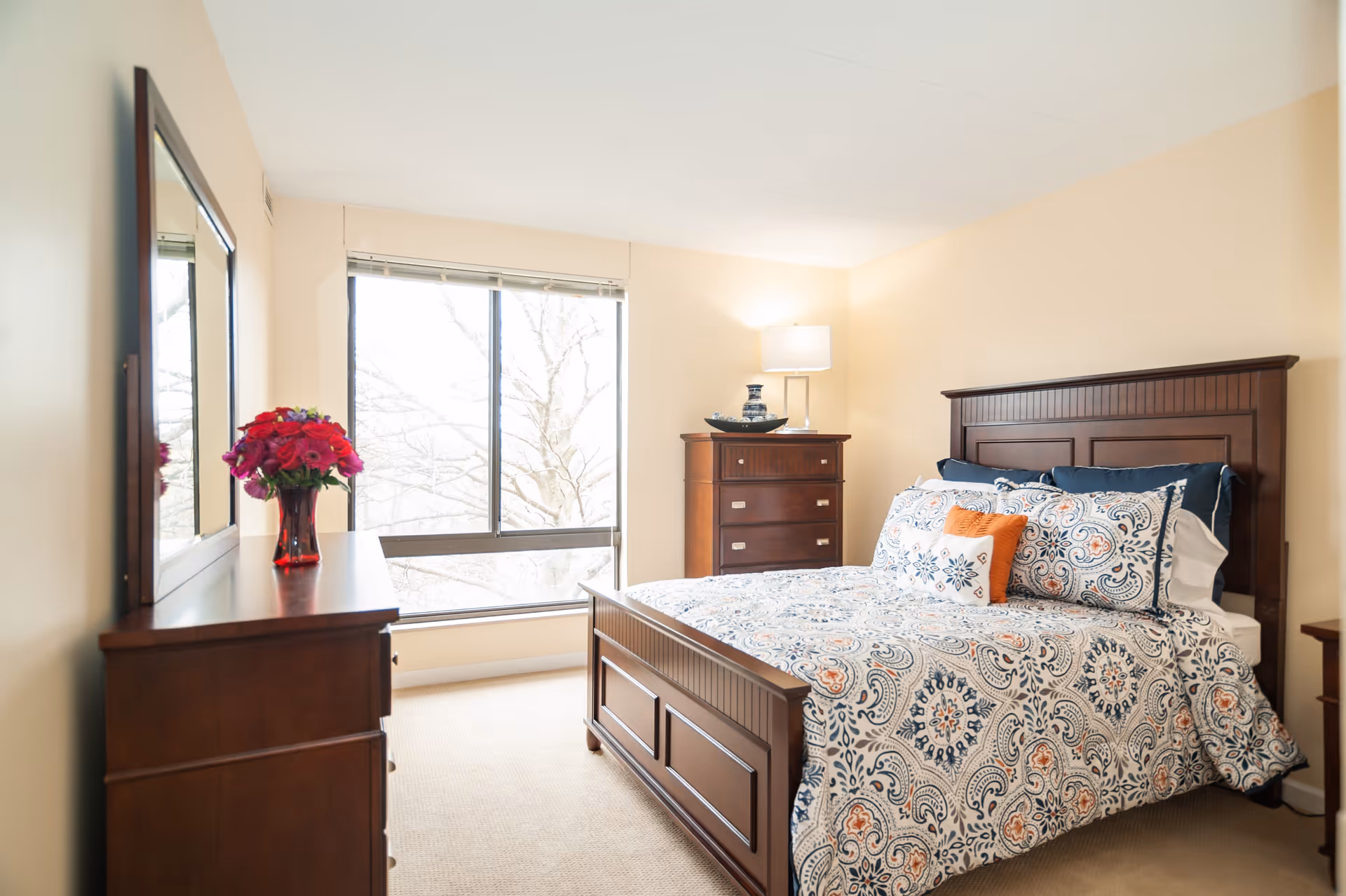 A bright bedroom with a large window letting in natural light. The room features a wooden bed with a patterned bedspread in blue, white, and orange tones, matching pillows, a wooden dresser with a vase of red flowers, a chest of drawers with a lamp and decorative items, and beige walls and carpet.