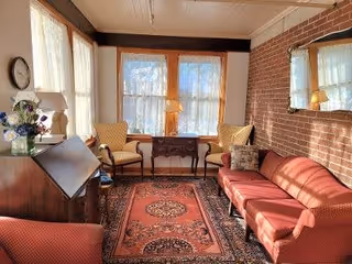 A cozy living room with a red sofa, two yellow armchairs, a wooden side table with a lamp between the chairs, a patterned rug on the floor, a wooden desk with a lamp and a flower vase, and a large mirror on a brick wall reflecting the room's light.