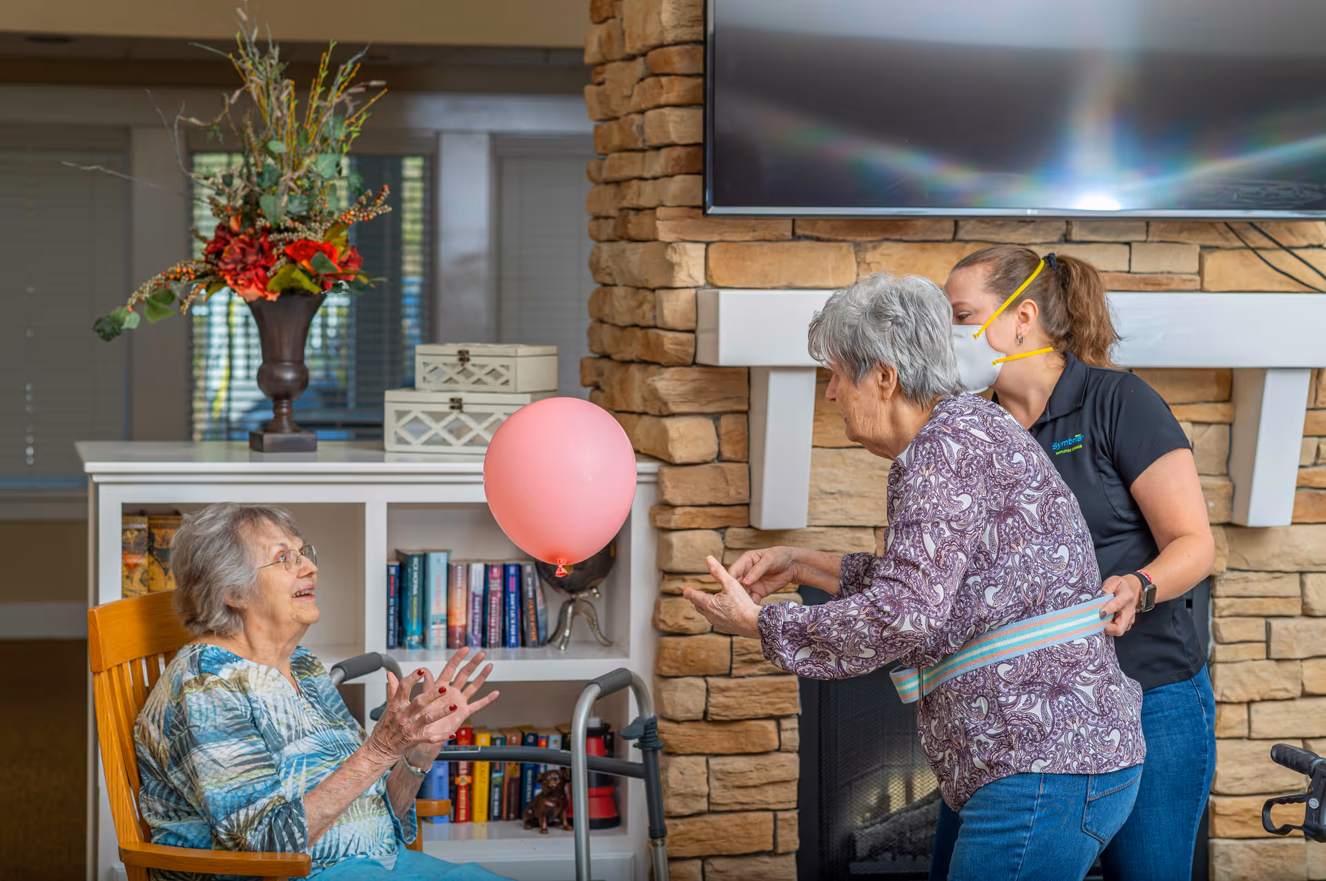 Two elderly women and a caregiver play with a pink balloon in a cozy senior living common room in front of a stone fireplace and bookshelf.