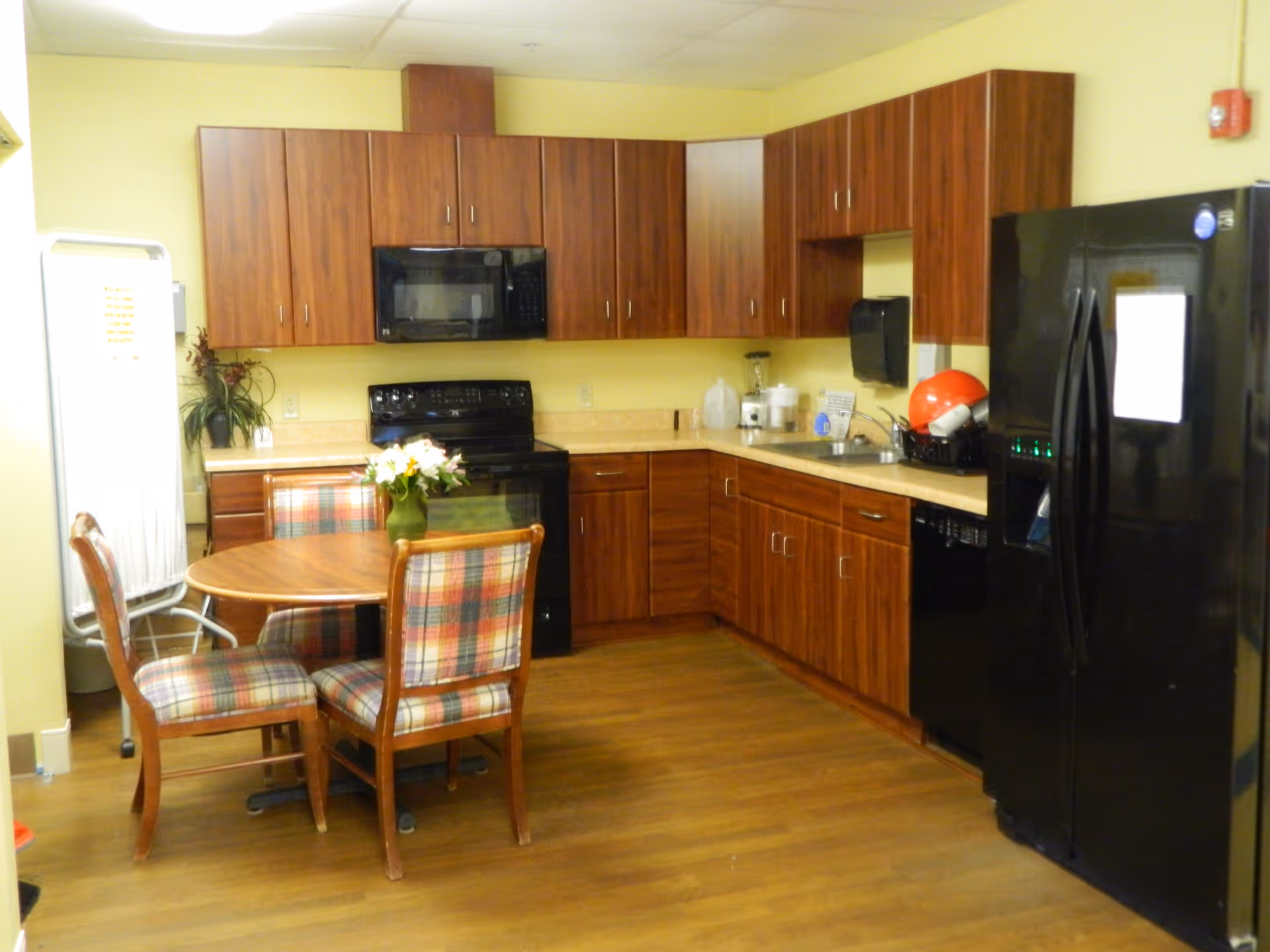 A kitchen area with wooden cabinets, a black refrigerator, black microwave, and black stove. There is a round wooden table with four plaid upholstered chairs in the center. The floor is wooden, and the walls are painted yellow. Various kitchen items and a vase with flowers are on the table and countertops.