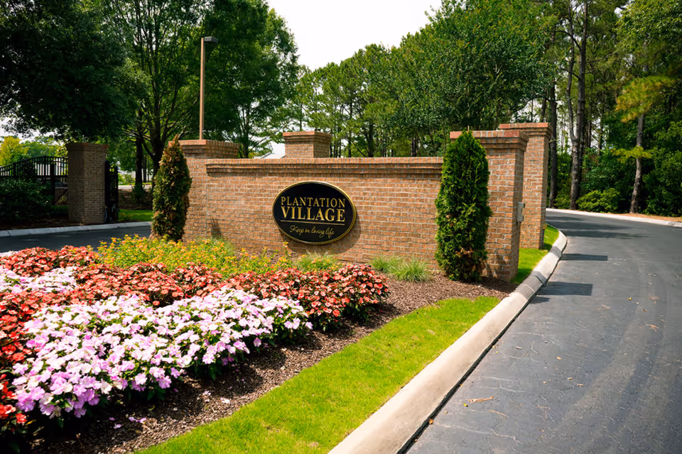 Brick entrance wall with a 'Plantation Village' sign, flower beds, and a tree-lined driveway.