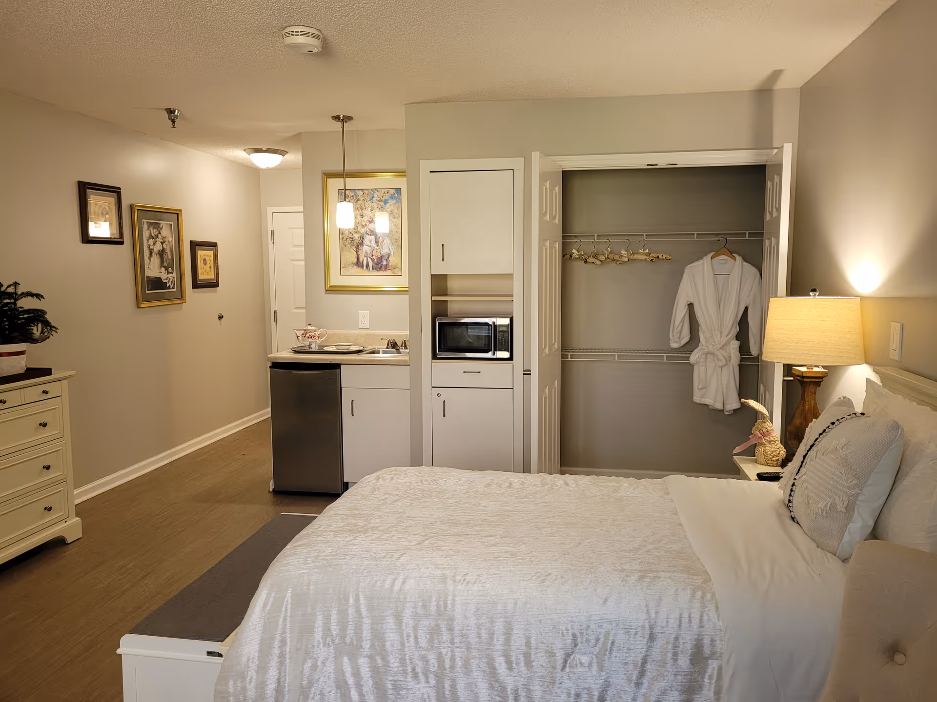 A cozy bedroom in a senior living facility featuring a neatly made bed with white bedding, a bedside table with a lamp and decorative items, an open closet with hangers and a white robe, a small kitchenette area with a mini fridge, microwave, and sink, and framed artwork on the walls.