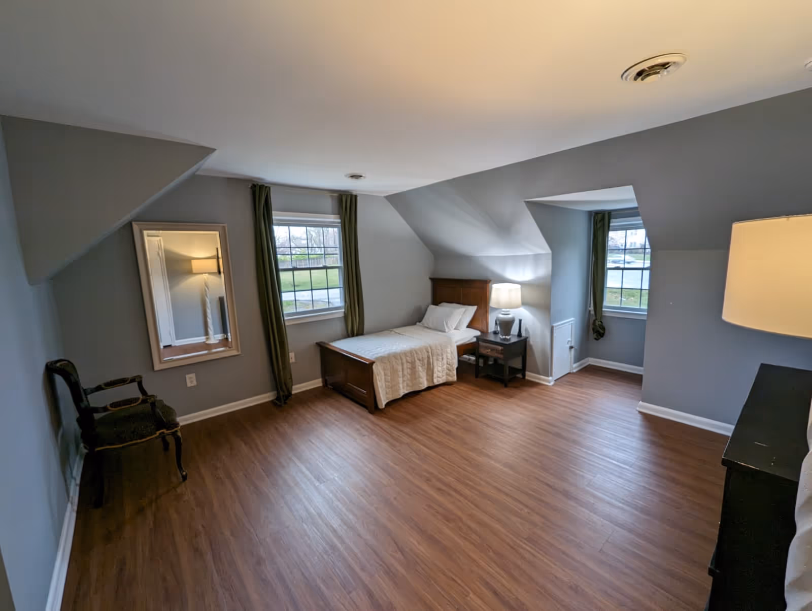 A spacious bedroom with a single bed, nightstand, lamp, and windows with curtains.