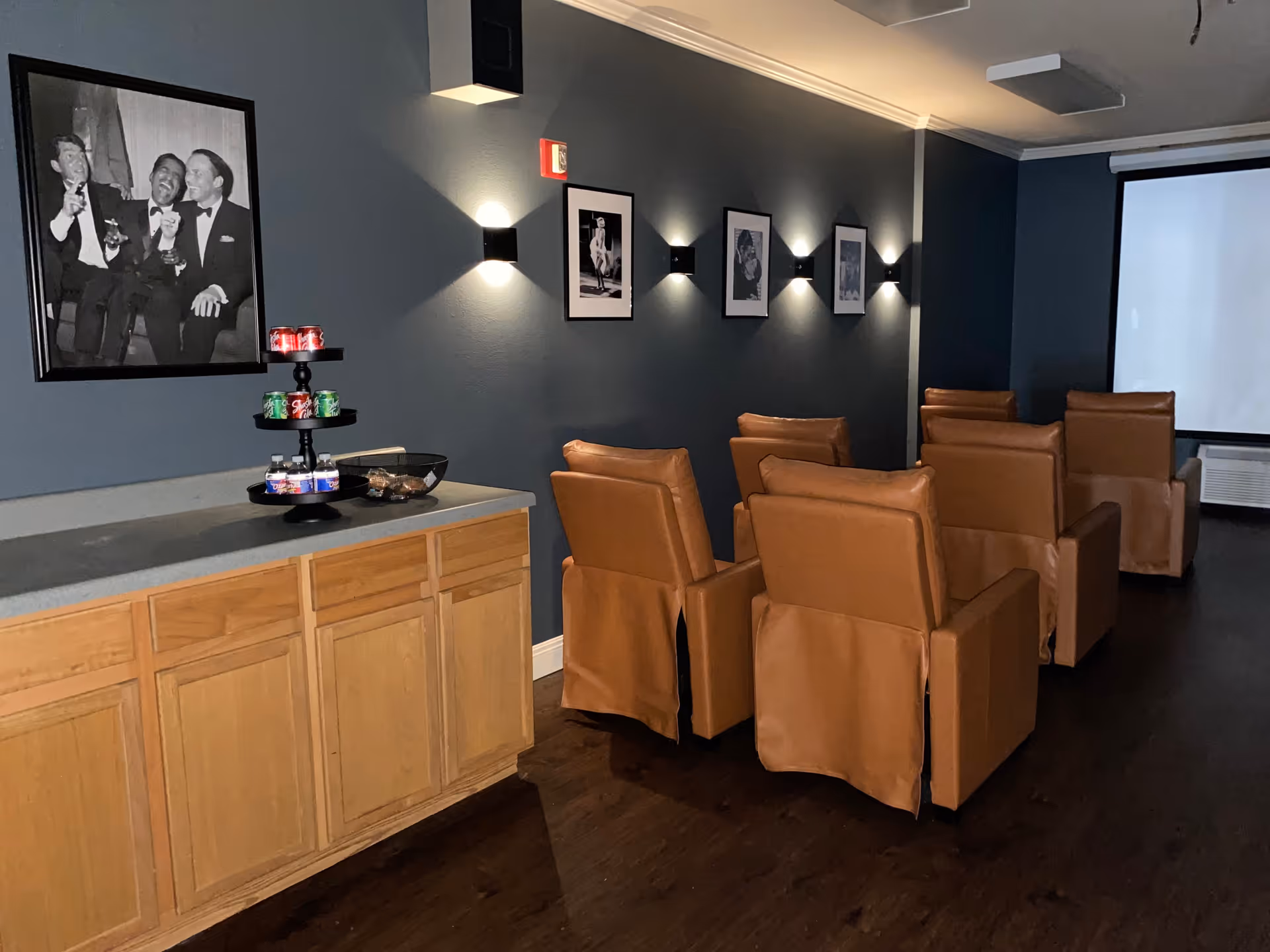 A small theater room with six brown leather recliner chairs arranged in two rows facing a large white projection screen. The walls are painted dark blue and decorated with black and white framed photographs. On the left side, there is a wooden cabinet with a tiered tray holding cans of soda and snacks.