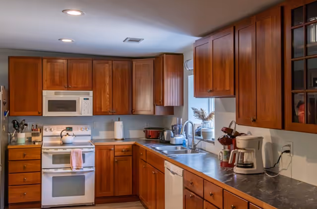 A kitchen with wooden cabinets, a white stove with a microwave above it, a dishwasher, a sink under a window, and various kitchen appliances on the countertops including a coffee maker and a blender.