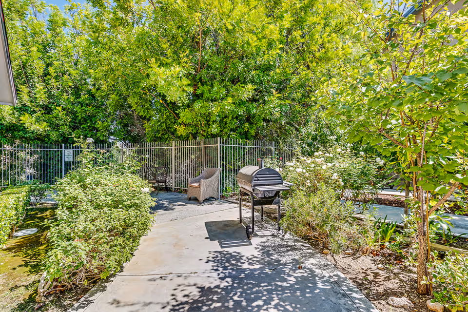 Outdoor garden area at Chino Hills Senior Living featuring a paved pathway, a charcoal grill, wicker chairs, lush green trees, bushes, and flowering plants under a bright blue sky.