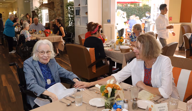 An elderly woman in a wheelchair and a middle-aged woman holding hands and smiling at each other while seated at a dining table in a busy assisted living community dining room. Other residents and staff are visible in the background, with tables set for a meal and a bright outdoor patio area beyond the open doors.