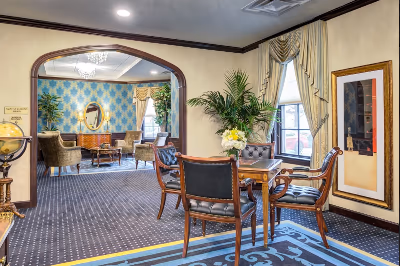 Elegant communal lounge with leather chairs around a small table, potted plants, and an archway revealing additional seating and patterned wallpaper.