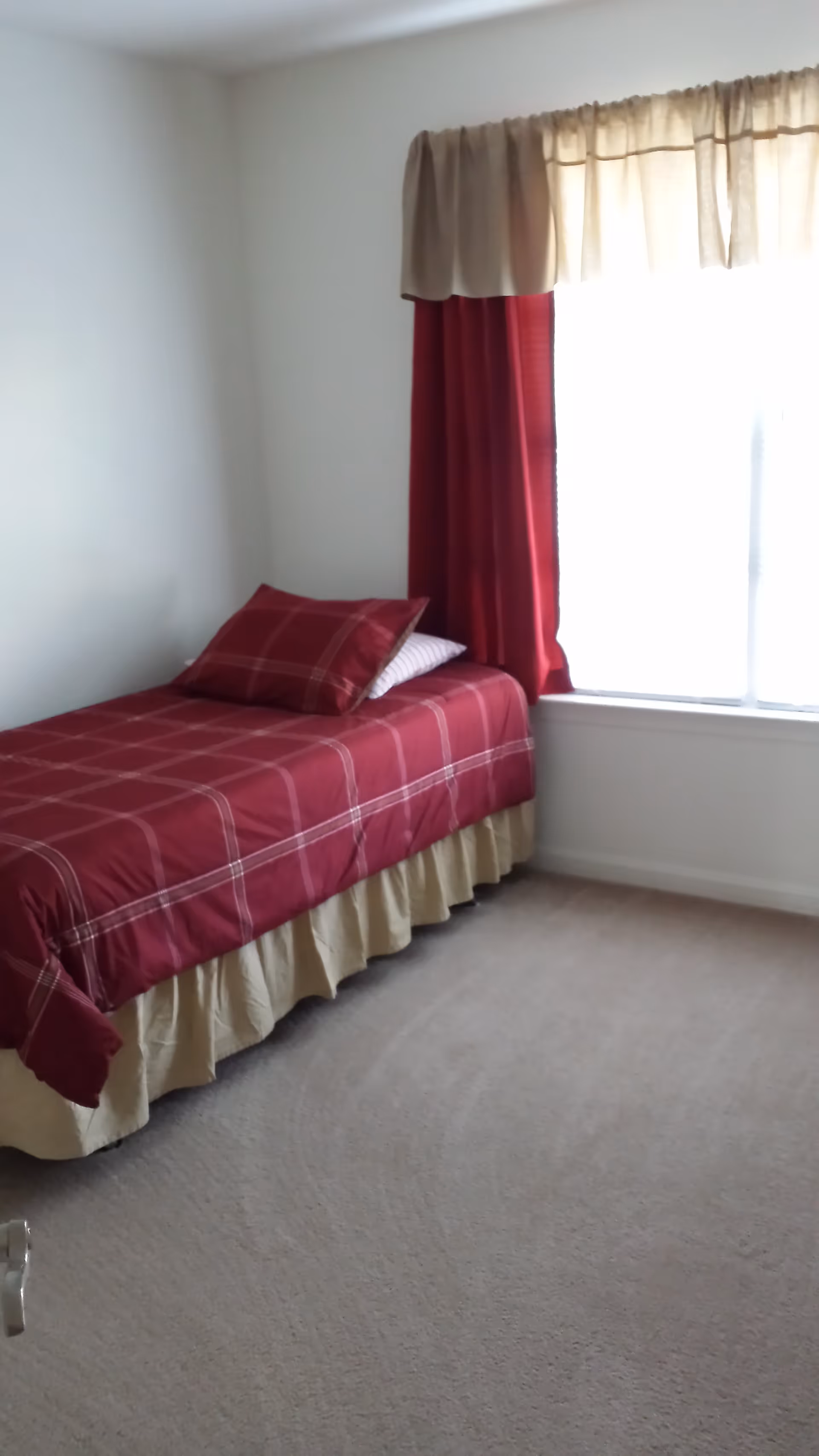 Small bedroom with a twin bed dressed in a red plaid bedspread and pillow next to a window with red curtains.