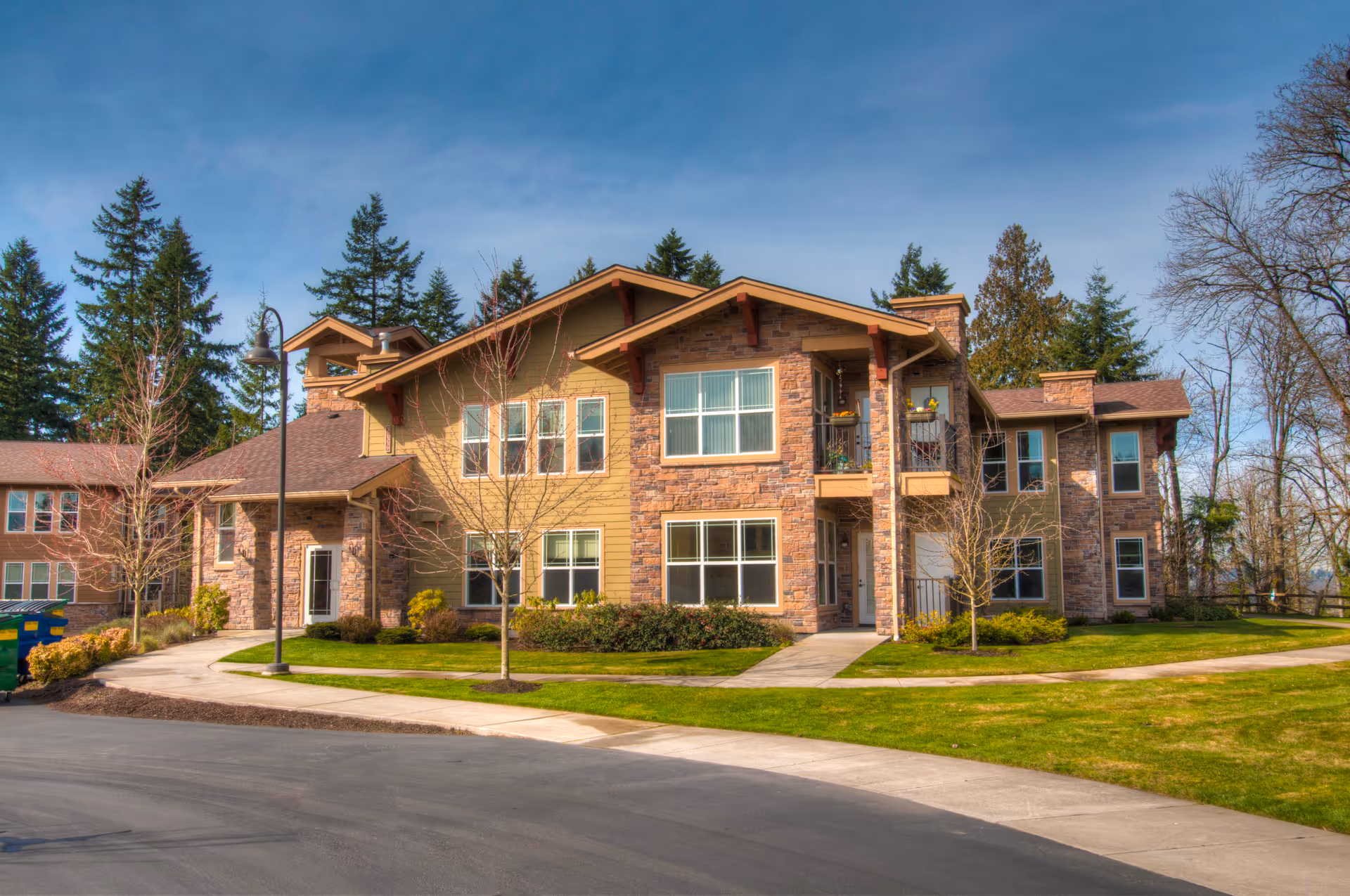 Exterior view of a two-story senior living facility building with stone and wood siding, surrounded by a well-maintained lawn, young trees, and a paved walkway under a clear blue sky.