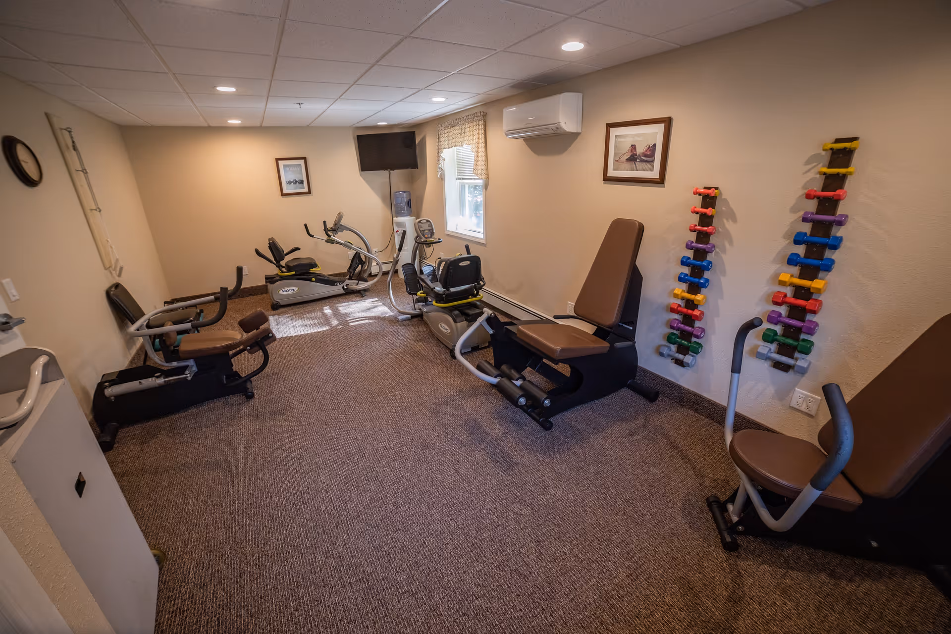 A small fitness room with exercise equipment including stationary bikes, a leg press machine, and a wall-mounted rack holding colorful dumbbells. The room has beige walls, a carpeted floor, a window with a curtain, a wall-mounted TV, and framed pictures on the walls.