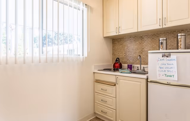 Small kitchen area with cream-colored cabinets, a white mini refrigerator with a handwritten note attached, a sink, a red kettle on the stove, and a window with vertical blinds letting in natural light.
