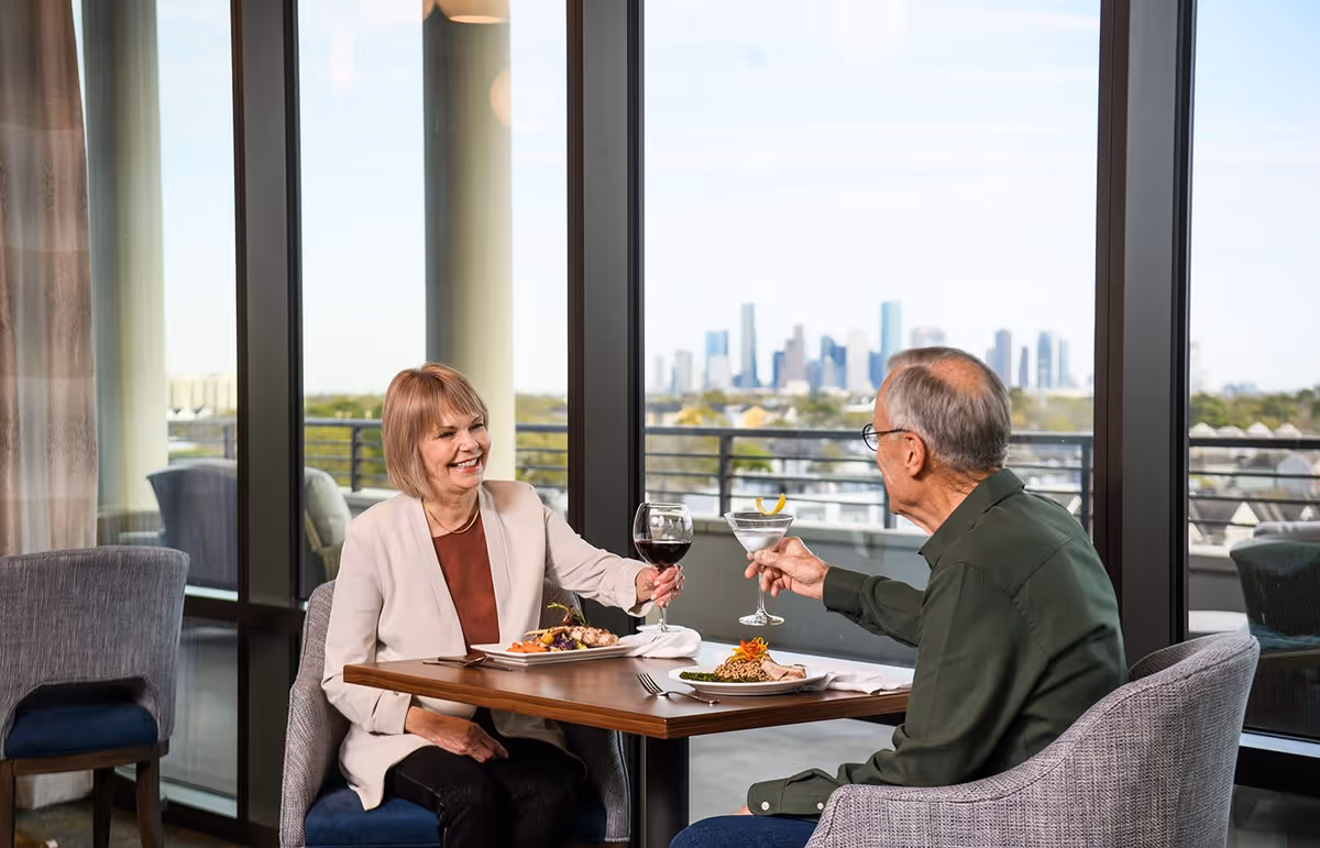 An elderly man and woman sitting at a dining table inside a room with large windows, enjoying a meal and toasting with drinks. The background shows a city skyline view through the windows.