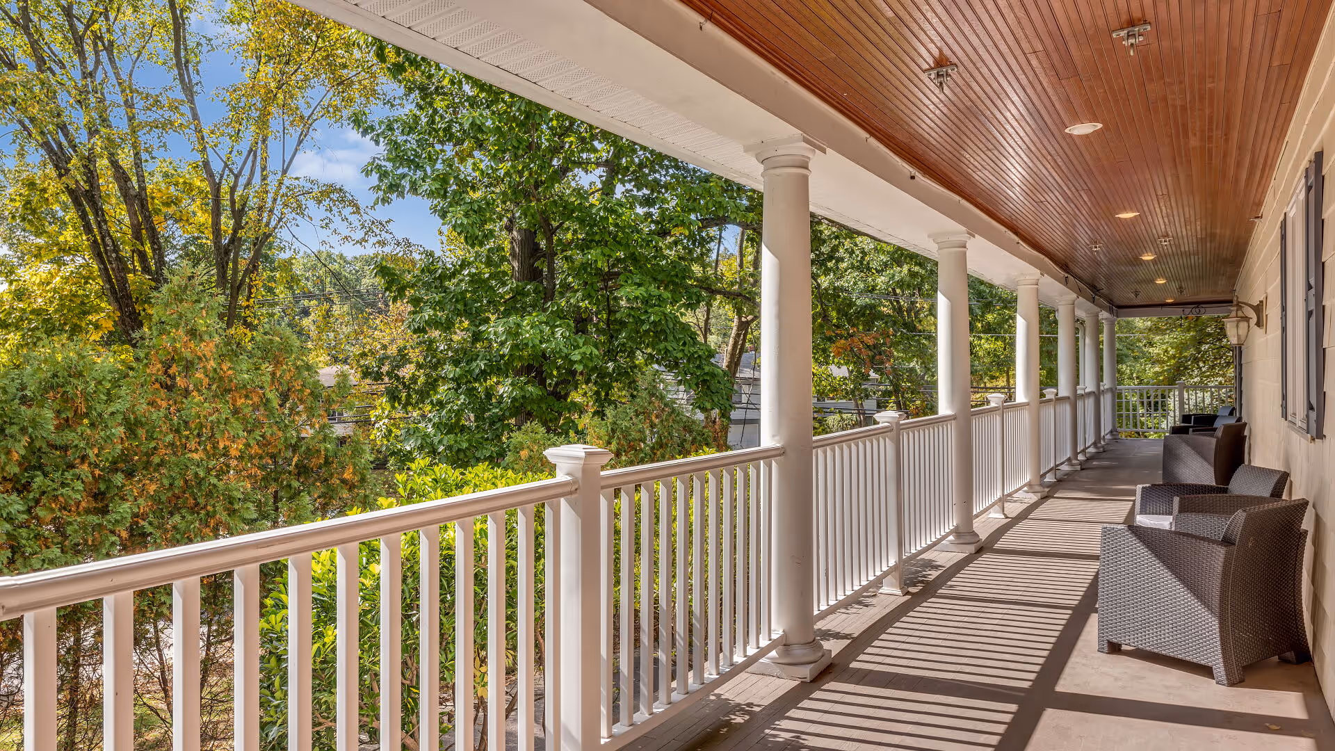 A long covered porch with white railings and columns, featuring several dark wicker chairs and a wooden ceiling. The porch overlooks a lush green area with trees under a blue sky.