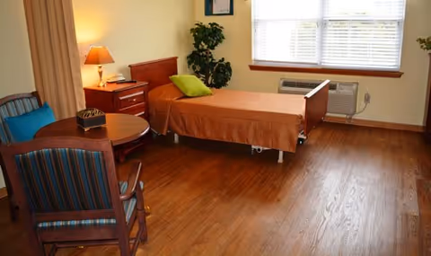 A senior living facility bedroom with a single bed covered in a brown bedspread and a green pillow. There is a wooden nightstand with a lamp and a phone next to the bed. A small round wooden table with two striped upholstered chairs is in the foreground. A large window with blinds and an air conditioning unit below it is on the far wall. A potted plant is placed near the bed, and the floor is wooden.