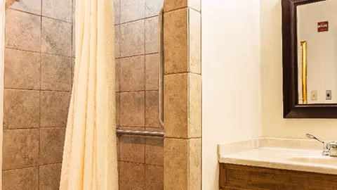 Bathroom with a tiled shower area featuring a beige shower curtain, a grab bar inside the shower, and a sink with a countertop and a mirror above it.