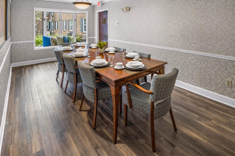 A dining room with a long wooden table set for eight people with white plates, bowls, cups, and glasses. The chairs around the table have patterned upholstery. There is a large window with a cushioned bench seat and colorful pillows, and the walls have a light patterned wallpaper with white trim. The floor is wood with a natural finish.