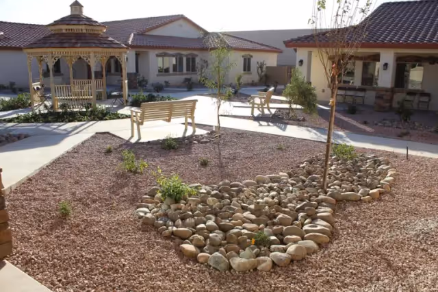 Outdoor courtyard area of a senior living facility with a wooden gazebo, benches, small trees, and landscaped rock garden beds surrounded by single-story buildings with tiled roofs.