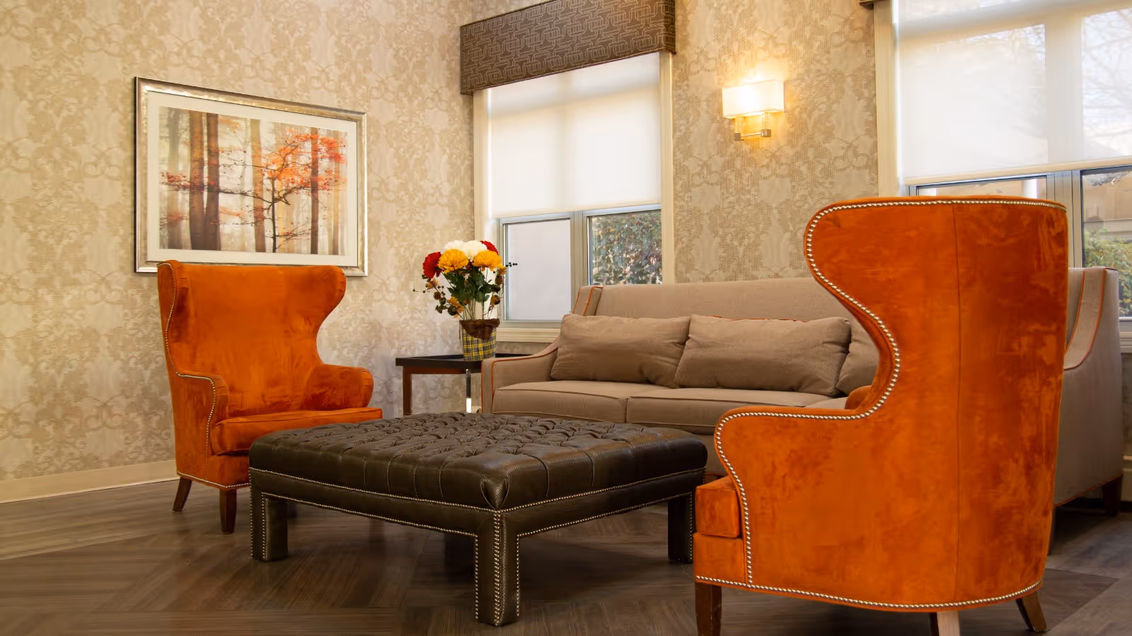 A cozy living room area with two bright orange wingback chairs facing a large, tufted black leather ottoman. Behind the ottoman is a beige sofa with several cushions. The walls have a patterned wallpaper, and there is a framed picture of trees with autumn leaves hanging on the wall. A small side table with a vase of colorful flowers is placed next to the sofa. Two windows with white roller shades allow natural light into the room.