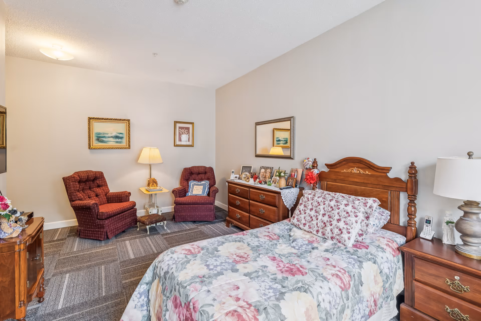 A cozy bedroom with a floral bedspread on a wooden bed, two red upholstered armchairs with a small table and lamp between them, a wooden dresser with framed photos and decorative items, and a nightstand with a lamp and phone. The walls are light-colored with framed artwork and a mirror above the dresser.