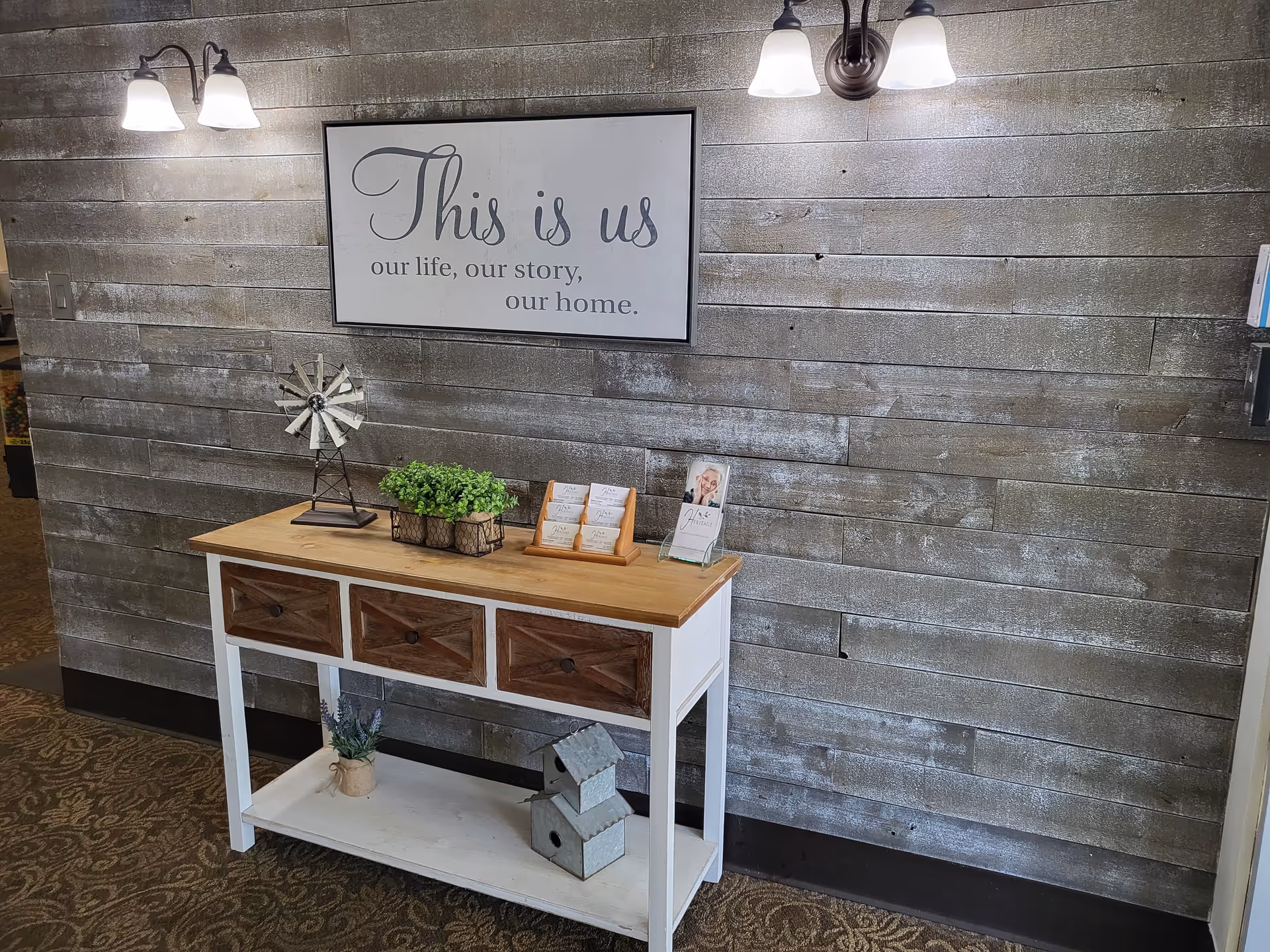 A rustic wooden console table with three drawers and a lower shelf is placed against a textured wooden plank wall. On the table are decorative items including a small windmill sculpture, a green plant in a wire basket, and two brochure holders. Above the table hangs a framed sign that reads 'This is us our life, our story, our home.' Two wall-mounted light fixtures with white shades illuminate the area.