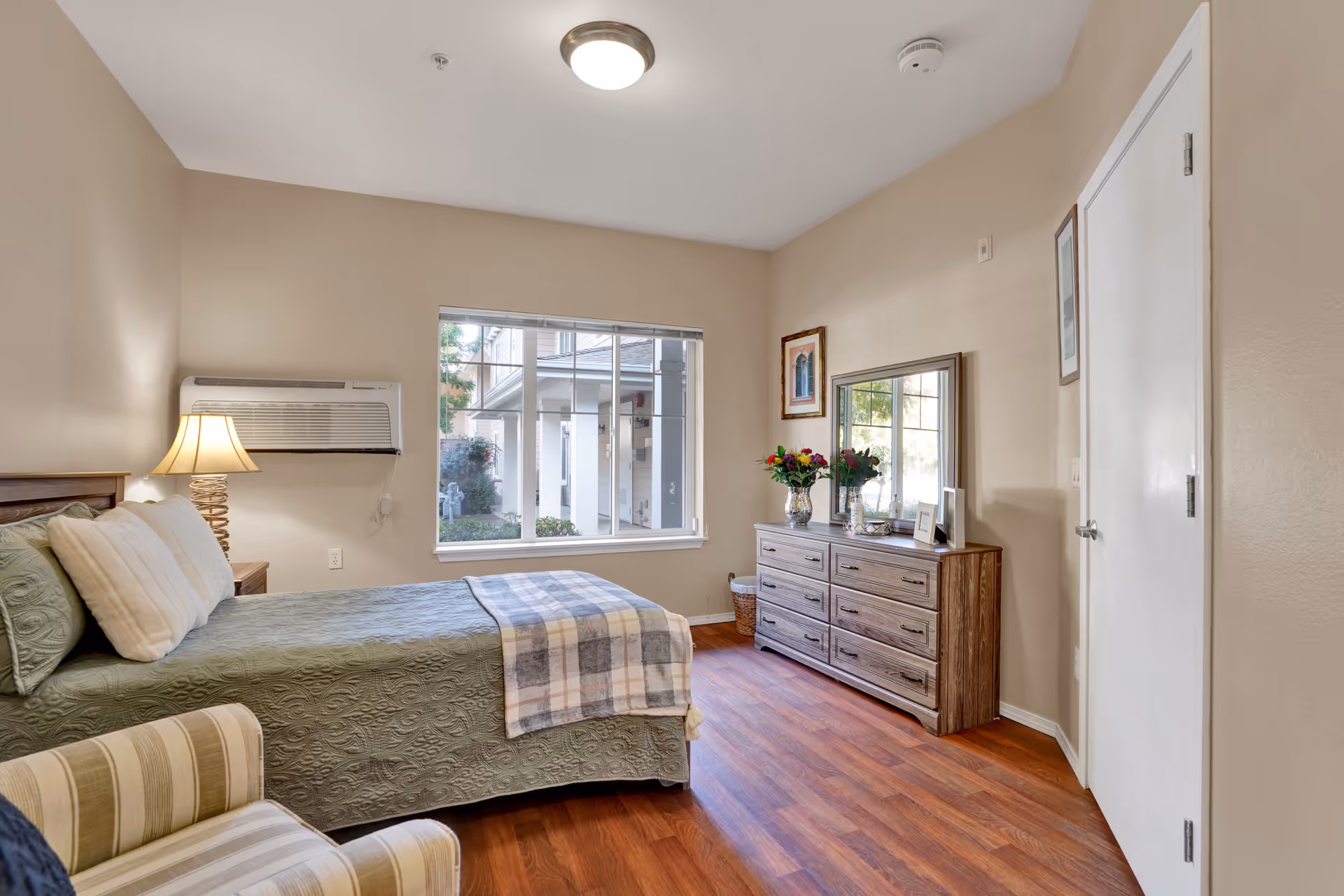 A cozy bedroom with a bed covered in a green quilt and white pillows, a wooden nightstand with a lamp, a striped armchair, a wooden dresser with a mirror, flowers in a vase, framed pictures on the wall, and a window showing an exterior view of the building.