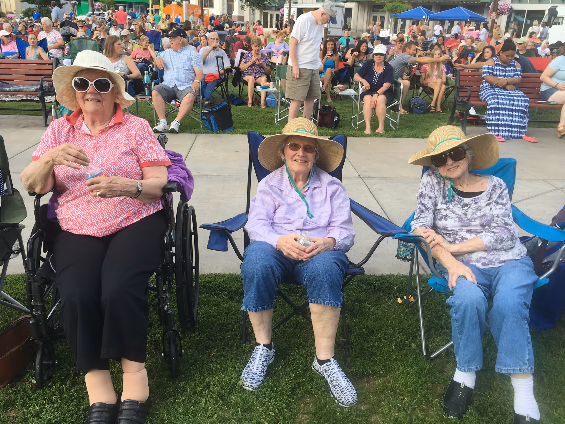 Three elderly women sitting outdoors on grass and pavement. The woman on the left is in a wheelchair wearing a white sunhat, sunglasses, a pink patterned shirt, and black pants. The two women on the right are seated in folding chairs, both wearing wide-brimmed sun hats and sunglasses. Behind them is a large crowd of people seated on benches and chairs, enjoying an outdoor event.
