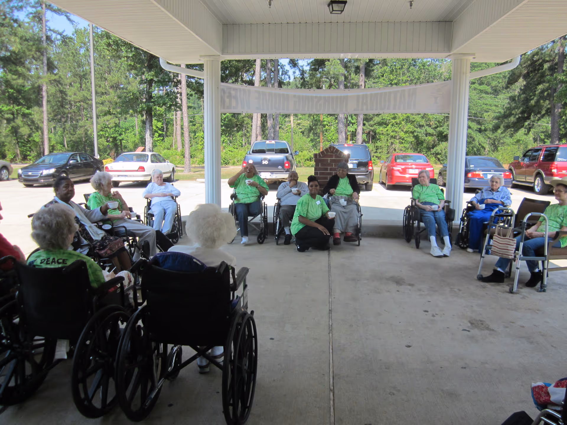 A group of elderly people, many in wheelchairs, sitting in a semi-circle under a covered outdoor area with trees and parked cars visible in the background. Some individuals are wearing green shirts, and one person is kneeling in front of the group.