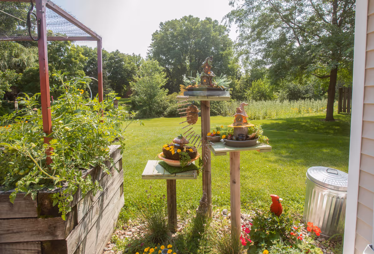 Sunlit backyard garden with a raised wooden planter, decorative plant stands holding potted flowers, a grassy lawn and trees beyond.