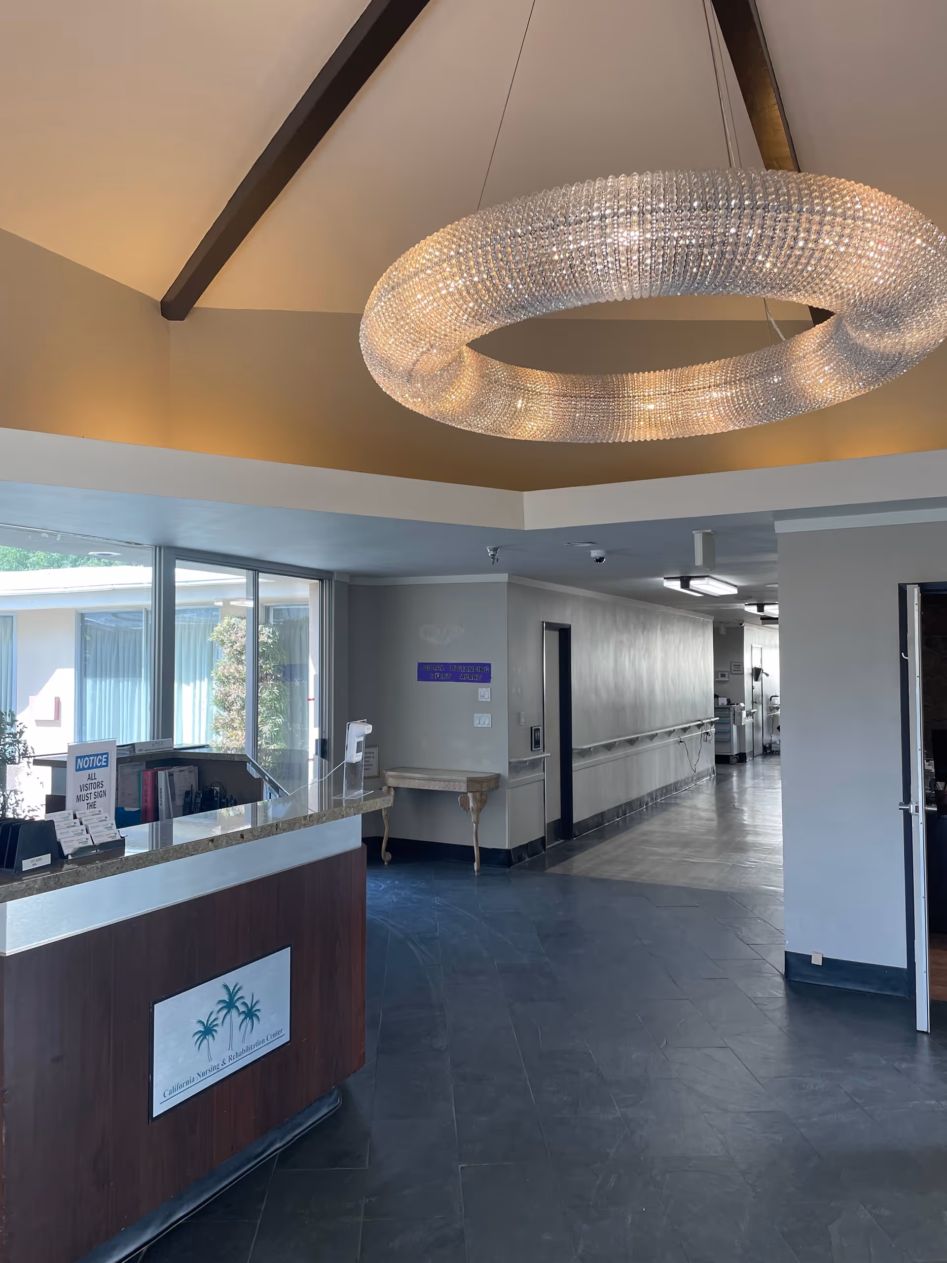 Reception area of California Nursing & Rehabilitation Center with a wooden front desk featuring a palm tree logo, a large circular chandelier hanging from a high ceiling with exposed beams, and a hallway leading to other rooms. There is a window with curtains and a small table against the wall.