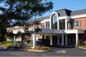 Exterior view of Mary Ann Morse Healthcare Center showing a two-story brick building with a covered entrance. A black car is parked under the entrance canopy, and there are trees and landscaping around the driveway.