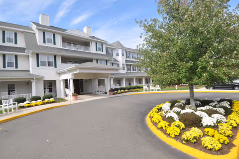 Front exterior view of a large, white senior living facility building with multiple windows, balconies, and a covered entrance. The driveway curves around a landscaped area with yellow and white flowers and a tree. White benches are placed near the entrance and along the building.