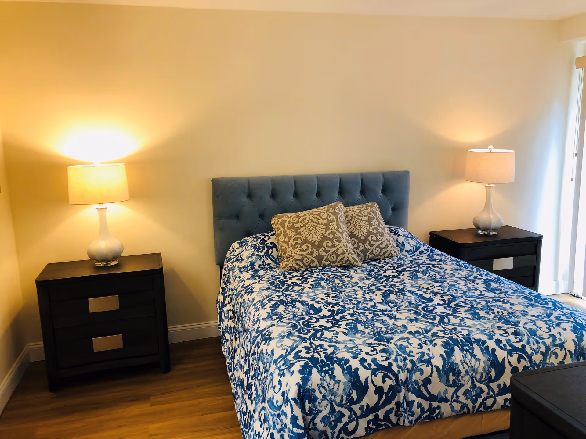 A tidy bedroom with a blue-patterned bed, tufted headboard, two bedside tables and lit lamps.