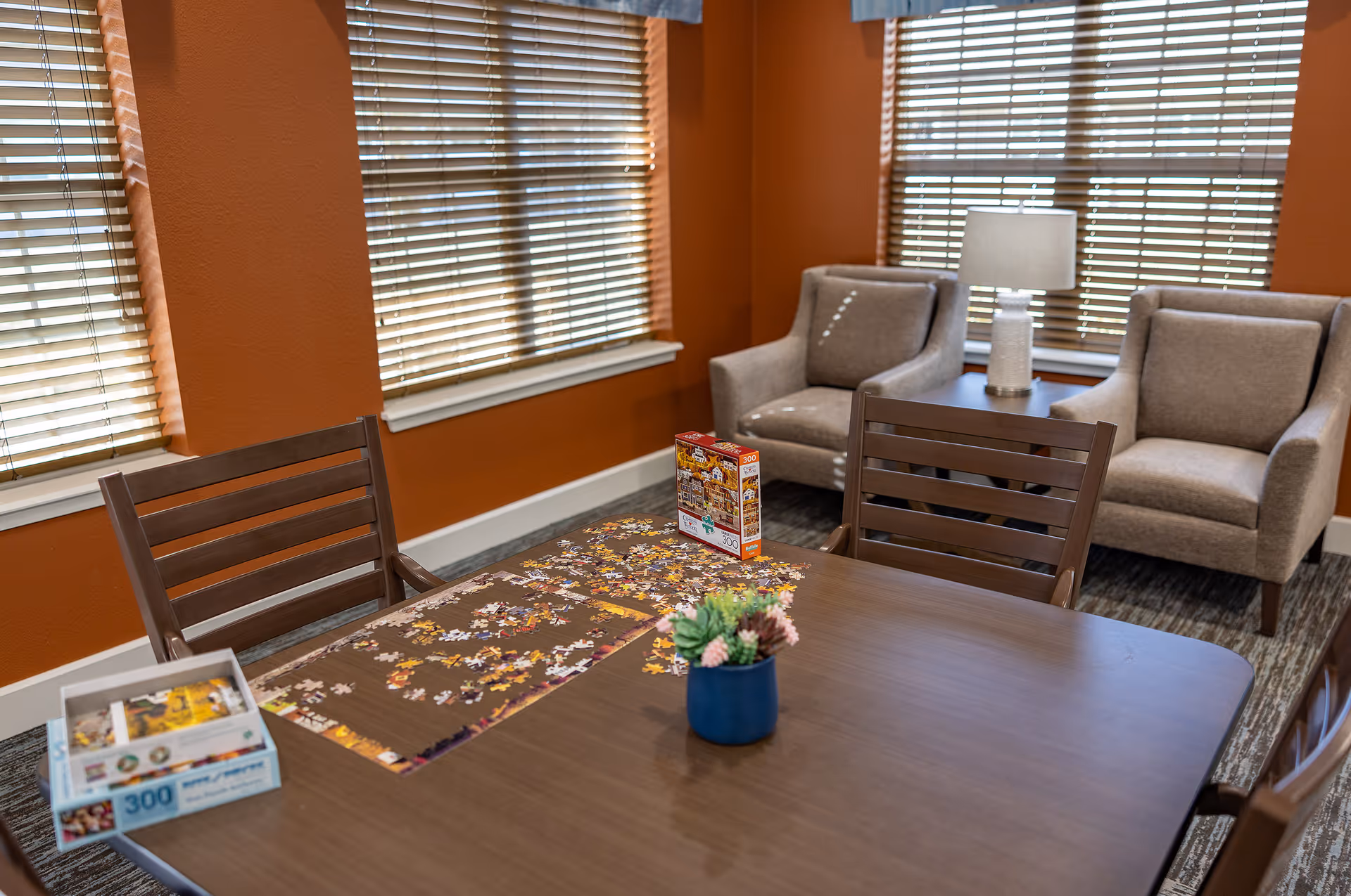 A cozy room with orange walls and large windows covered with blinds. There is a wooden table in the center with a partially completed 300-piece jigsaw puzzle and puzzle boxes on it. Two wooden chairs are positioned around the table. In the background, there are two beige armchairs with a small side table and a lamp between them.