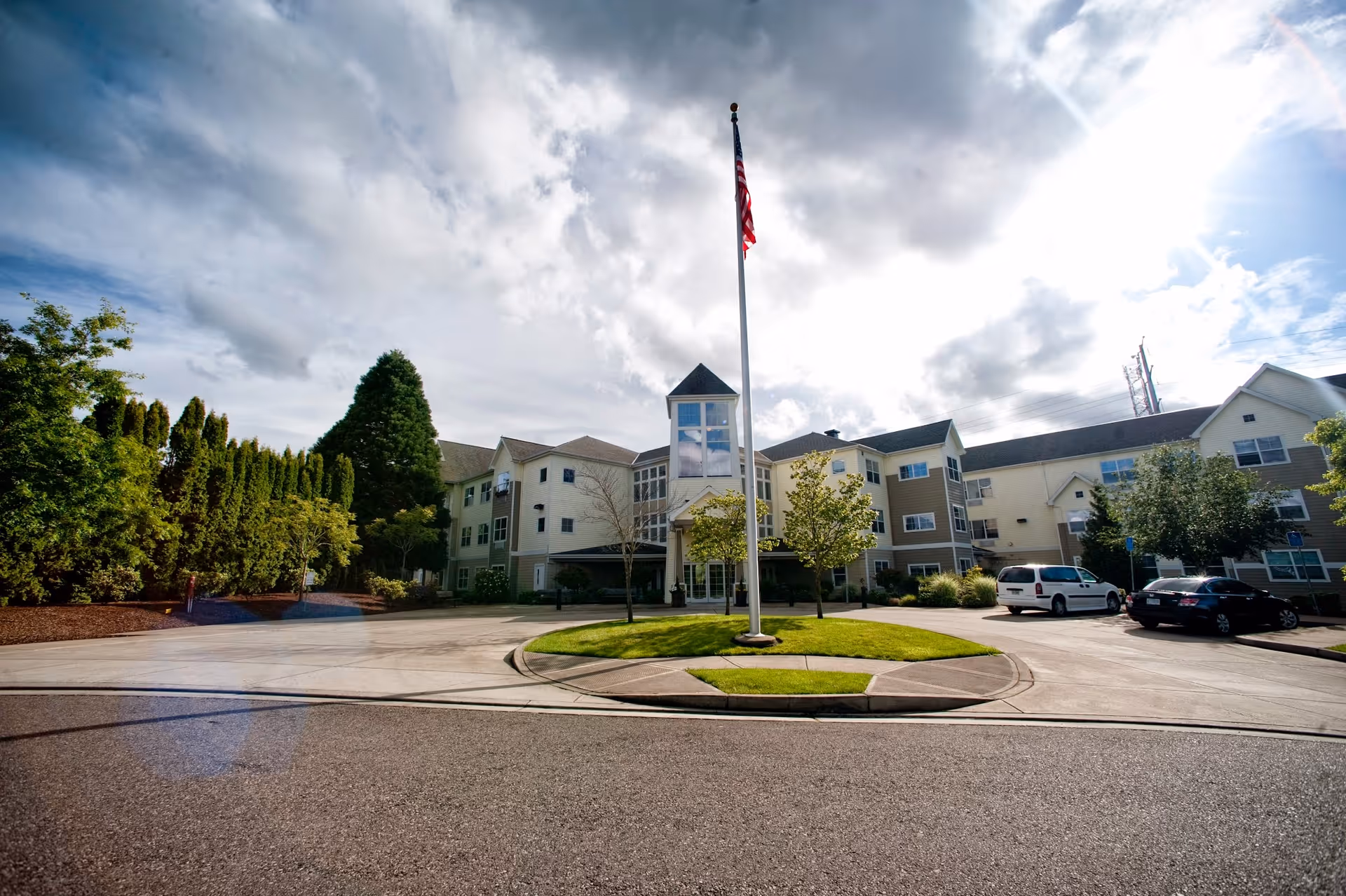 Exterior view of a multi-story senior living facility with a circular driveway and a flagpole with an American flag in the center. The building is surrounded by trees and parked cars under a partly cloudy sky.