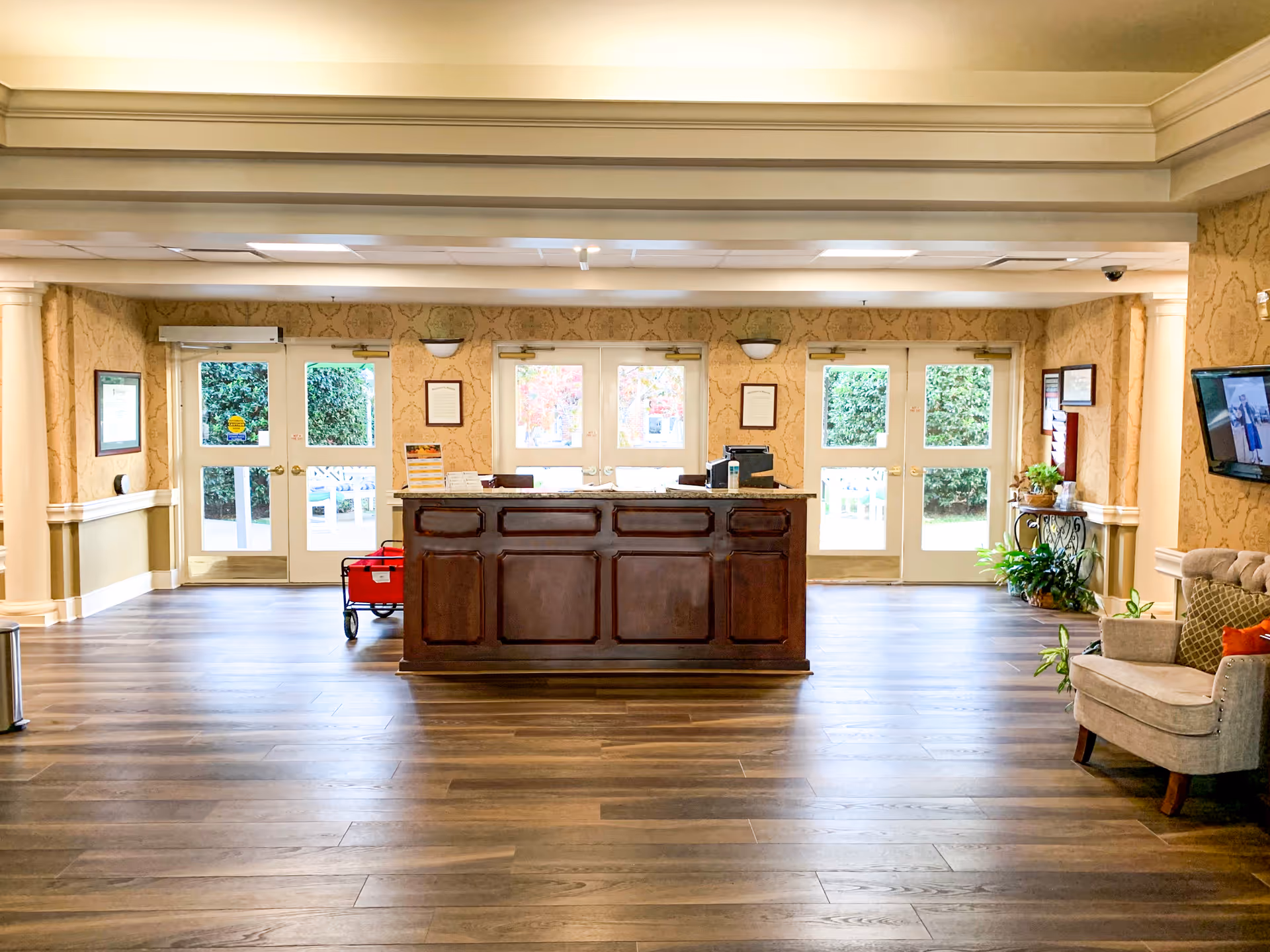 Reception area of a senior living facility with a wooden front desk in the center, light-colored walls with decorative molding, multiple glass doors letting in natural light, a beige armchair with cushions on the right, a wall-mounted TV, and some potted plants near the walls.