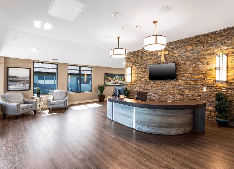 A bright and spacious reception area with a curved wooden desk in front of a stone accent wall. The wall features a mounted flat-screen TV and a decorative cross. Two modern pendant lights hang from the ceiling, and two comfortable armchairs with a small table are placed near large windows letting in natural light. There are also two potted plants and framed artwork on the walls.