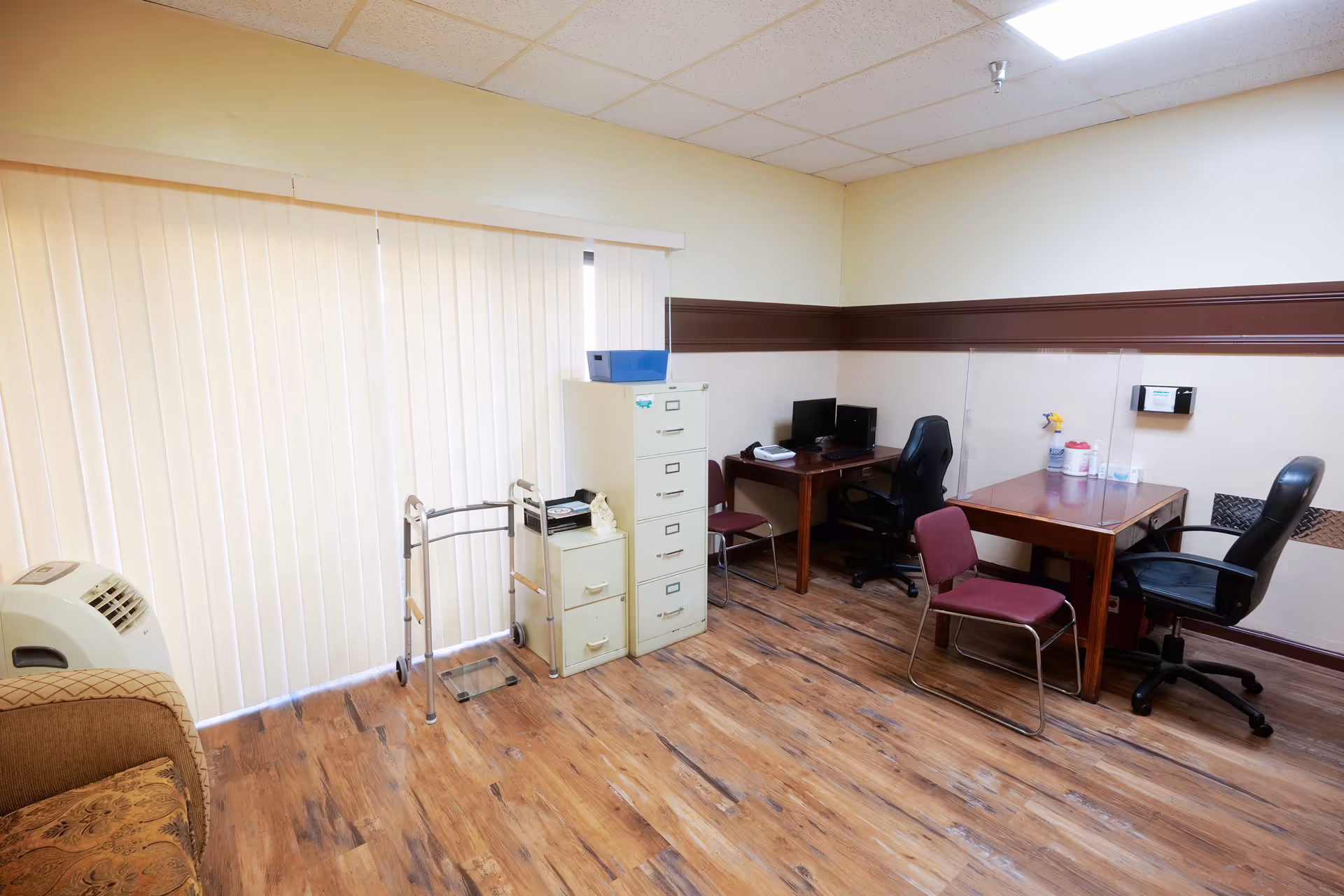 An office space with wooden flooring and beige walls featuring a vertical blind-covered window. The room contains a beige filing cabinet, a walker, two desks with office chairs, a maroon chair, a computer monitor, and cleaning supplies on one desk. A couch is partially visible on the left side.