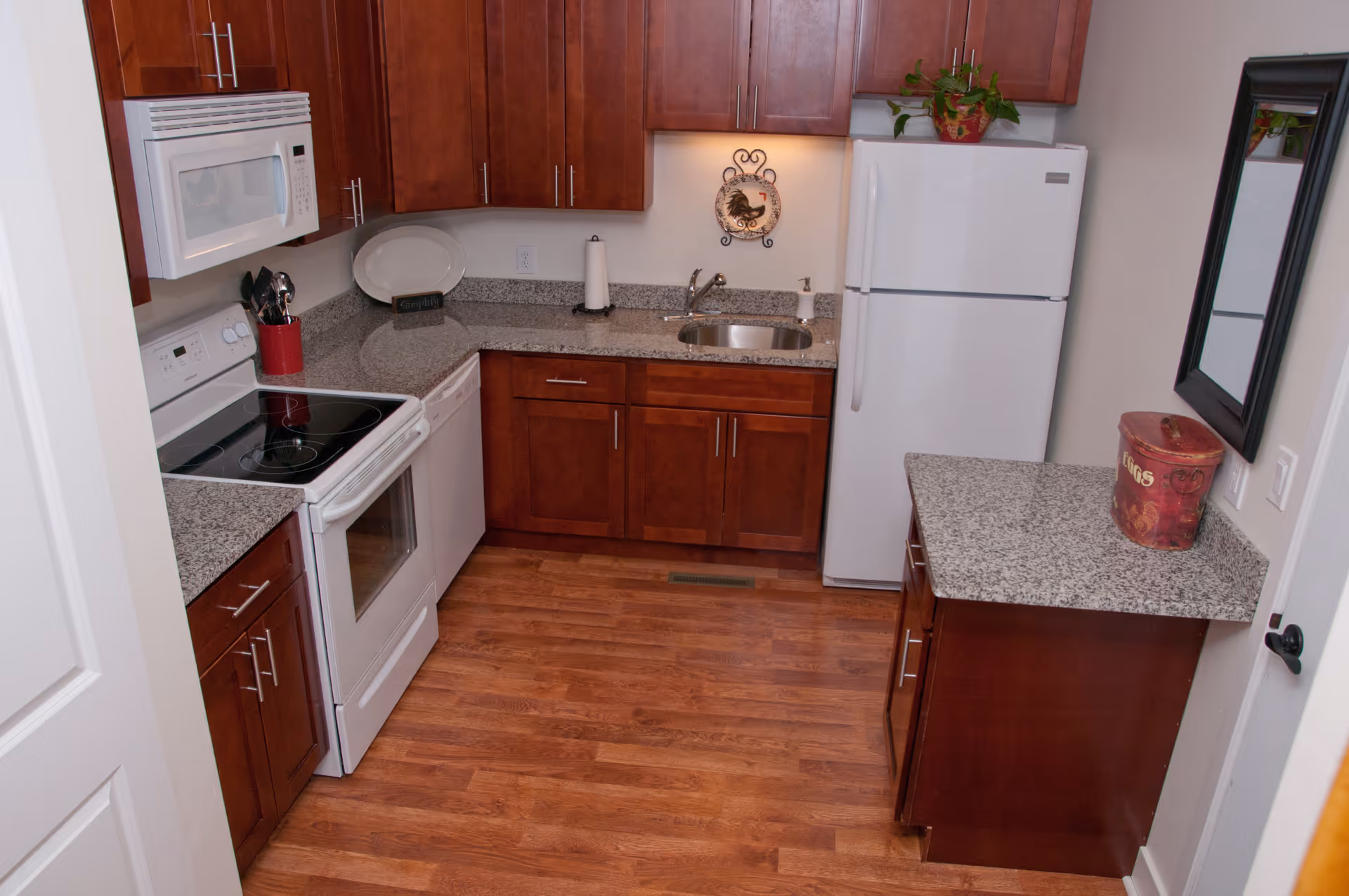 Compact kitchen with cherry wood cabinets, granite countertops, white stove, microwave and refrigerator, and wood-look flooring.