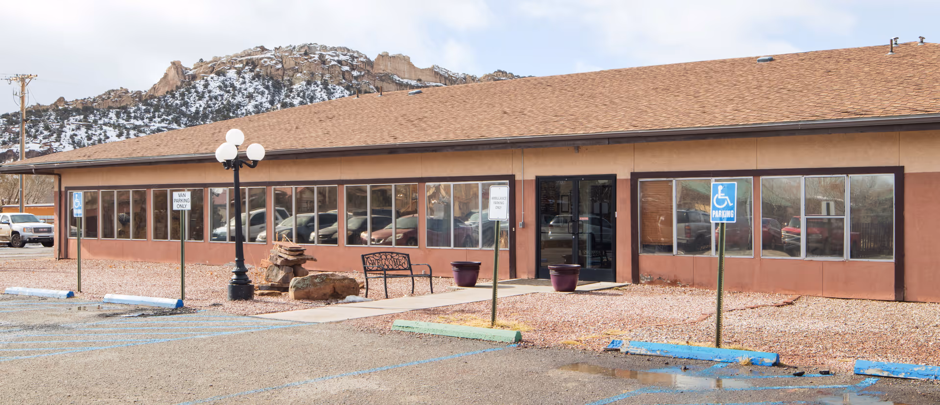 Exterior view of a single-story building with large windows and a brown roof, surrounded by a parking lot with handicap parking spaces. There is a bench and some decorative rocks near the entrance, with a mountainous landscape in the background.