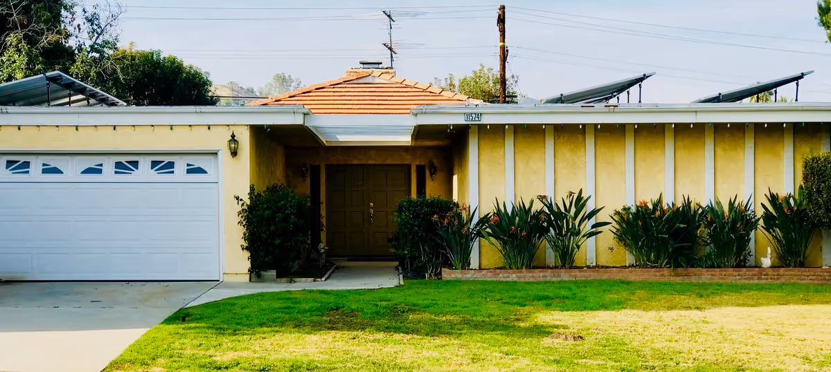 Front exterior view of a single-story house with a yellow facade, a white garage door on the left, a central entrance with double doors, and a well-maintained lawn with plants and shrubs in front.