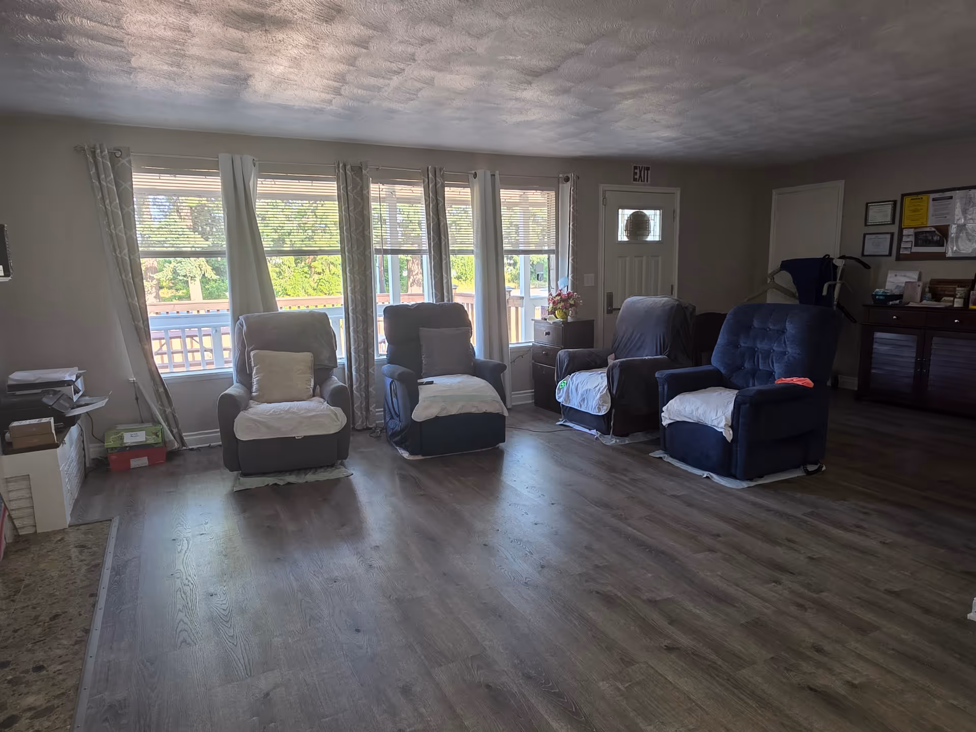 A living room area with four cushioned recliner chairs arranged in a semi-circle facing the windows. The room has large windows with curtains and blinds, letting in natural light. There is a door with an exit sign above it, a side table with flowers, and a cabinet with various items on top. The floor is wood laminate, and the walls are painted light gray.