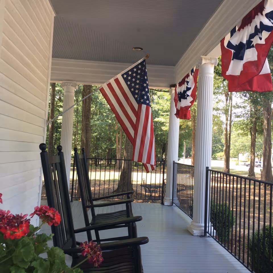 Covered front porch with black rocking chairs, white columns, an American flag and patriotic bunting overlooking a wooded yard.