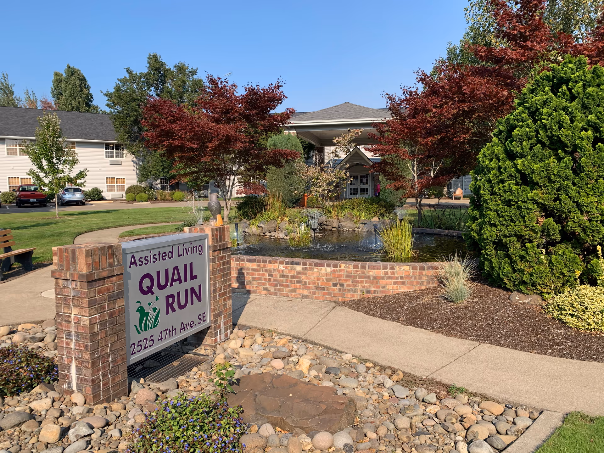 Outdoor view of an assisted living facility named Quail Run with a brick sign displaying the name and address. The scene includes a small pond with fountains, landscaped greenery, trees with red and green leaves, a paved walkway, and a building in the background under a clear blue sky.