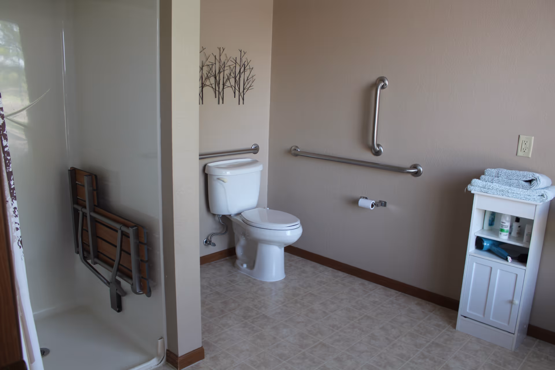 A bathroom with a white toilet, grab bars on the walls, a small white cabinet with towels and toiletries, and a shower area with a fold-down wooden seat attached to the wall.